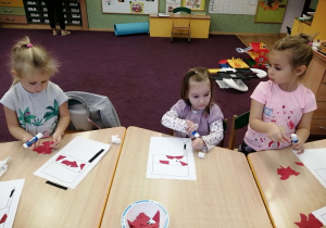 Dzieci siedzą przy stolikach, wyklejają białym i czerwonym papierem kontur flagi.